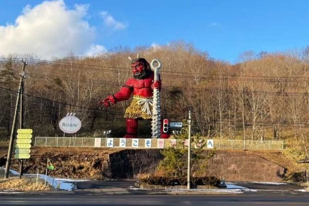 道路脇の小高い場所に立つ、巨大な赤い鬼の像。像は金棒を持っていて、看板には「ようこそ」と書かれている。