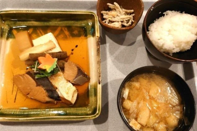 魚の煮付けやご飯、味噌汁などが並んだ美味しそうな和定食。リゾートバイトの賄いとして、温かみのある手作りの食事が提供され、食費を節約できるという魅力を伝えます。