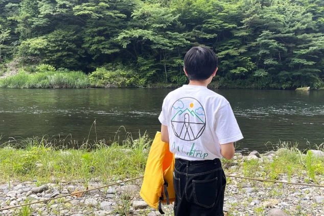 ワクトリのロゴ入りTシャツを着た人物が、鬼怒川の川辺に立つ後ろ姿。リゾートバイトの勤務地で、川のアクティビティを満喫できる様子を伝える画像。