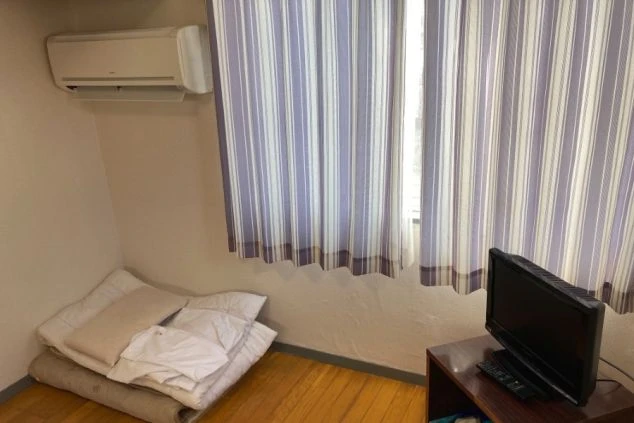 寮の個室。ベッド、テレビ、エアコン、カーテンが映る部屋の様子。