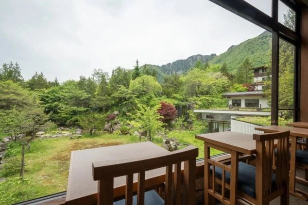 縁側にあるテーブルと椅子の手前から、窓の外に広がる緑豊かな庭園と山々を眺める旅館のロビーの画像。