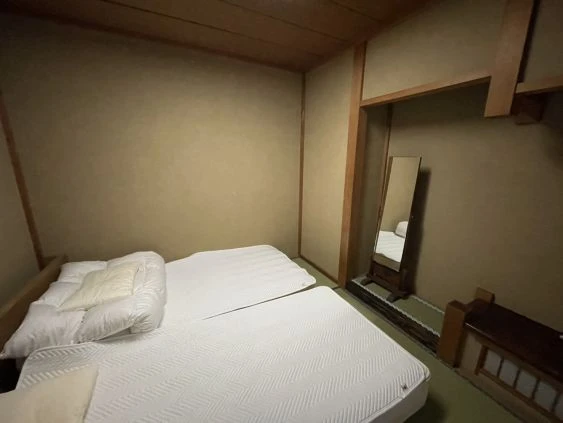 畳の部屋に、ベッドが2つ並べて置かれている旅館の客室寮の画像。部屋の奥には姿見と荷物を置くスペースが写っている画像。