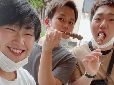 リゾートバイトの従業員が休日に観光を楽しむ様子を写した画像。串に刺さった食べ物を頬張る、男性3人の楽しそうな自撮り画像。