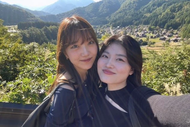 緑豊かな山々を背景に、楽しそうに寄り添って自撮りするリゾートバイトの女性2人組の画像。