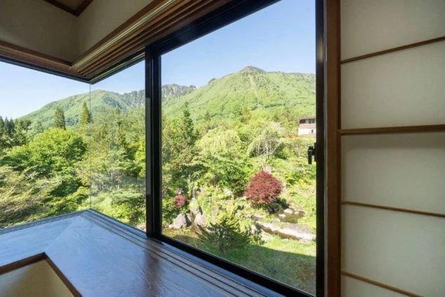 窓の外に新緑の山々が広がる、旅館の客室からの眺めの画像。窓の右側には障子が見え、左下には窓に沿って作られた木製の座卓が写っている画像。