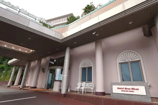 ピンクベージュの壁面に丸窓があしらわれたモダンな建物と、周囲を彩る豊かな木々。