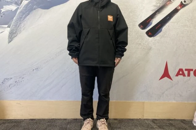 黒いジャケットとズボンを着用した人物。ジャケットの胸にはオレンジ色のロゴが入っている。背景にはスキーやスノーボードのポスターがある。