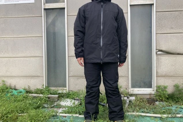 黒いスキーウェアを着用した男性。黒いジャケットとパンツを身につけ、建物の前で立っている。