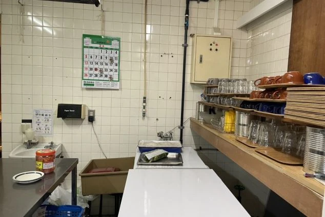 業務用厨房の一部。壁は白いタイル張りで、シンク、作業台、コップや皿が置かれた棚がある。