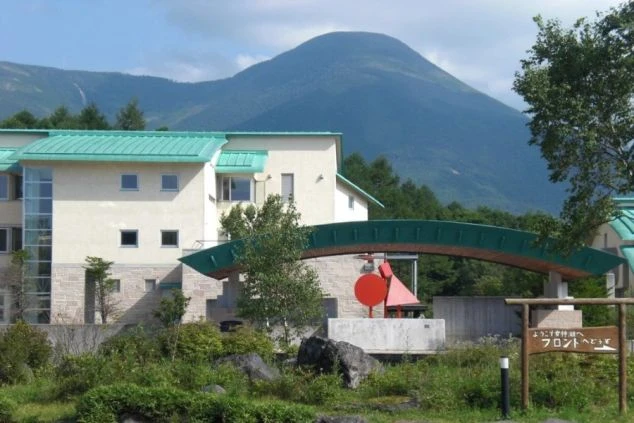 緑の屋根をした白い建物。建物の前には、赤いオブジェと、木製の看板が立っている。背景には山が見える。