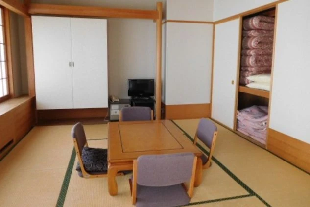 畳敷きの部屋。中央には座卓と4脚の座椅子が置かれている。部屋の奥には、布団が収納された押し入れとテレビが見える。