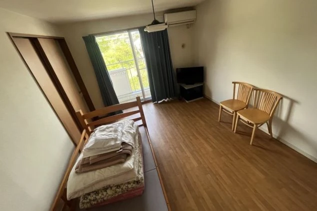 リゾートバイトの個室寮の室内。シングルベッド、テレビ、椅子が備え付けられ、窓からは光が差し込む明るい部屋の画像。