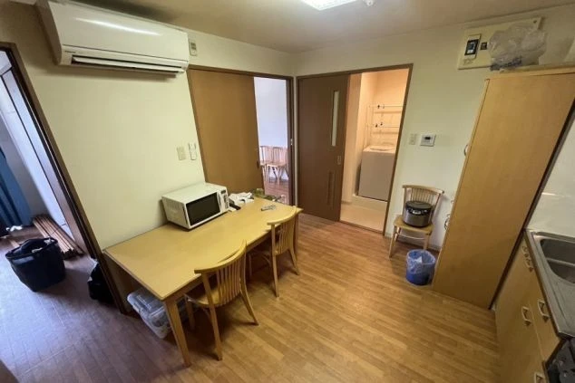 リゾートバイトの個室寮のダイニングスペース。テーブルと椅子、電子レンジが置かれ、奥には洗濯機や炊飯器が見える、自炊や食事ができる設備の寮の画像。