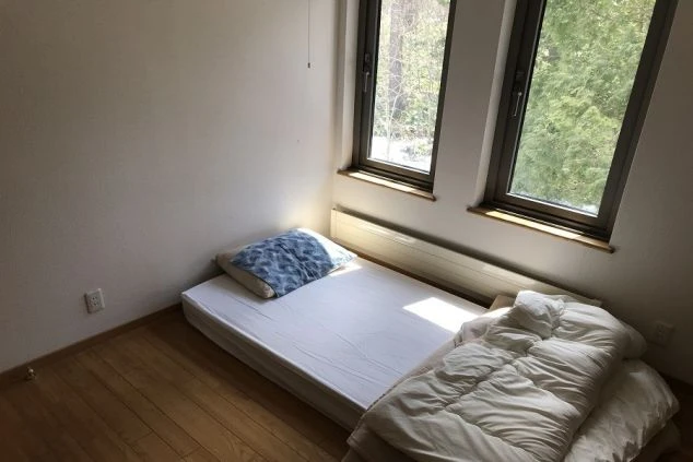 二つの窓から光が差し込む寮の部屋。床は木製で、布団が敷かれている。