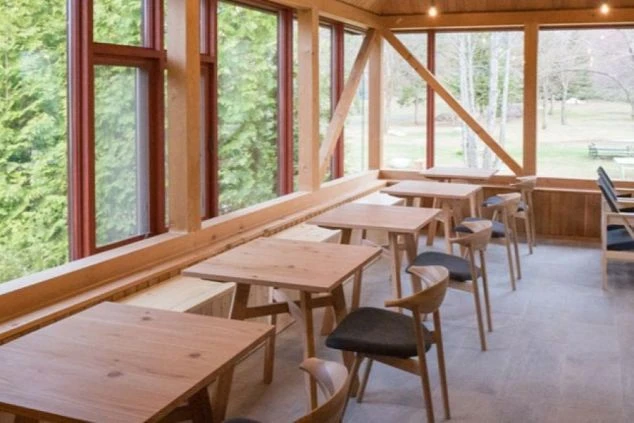 大きな窓から緑の木々が見えるカフェ。木製のテーブルと椅子が規則正しく並んでいる。