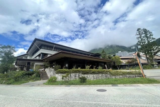 旅館の建物の外観を写した画像。背景には山がそびえており、空には雲が浮かんでいる画像