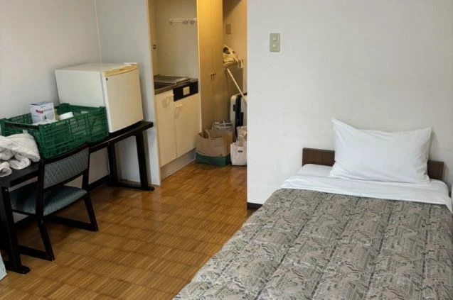 リゾートバイトの寮の部屋の画像。ベッドとデスク、冷蔵庫が置かれ、奥にはキッチンが見える。