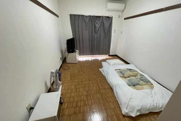 リゾートバイトの寮の個室の画像。布団が敷かれ、エアコン、テレビ、冷蔵庫、電子レンジ、掃除機が備え付けられている。