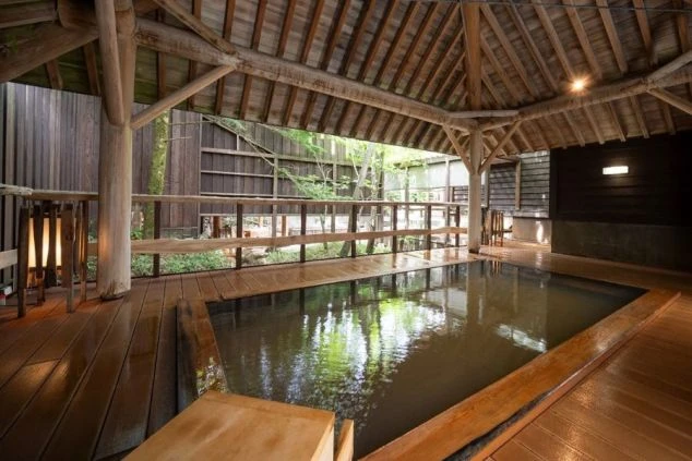 木造の屋根付きの、広々とした旅館の露天風呂の画像。浴槽には温泉が満たされており、周りは木々の緑に囲まれている画像。