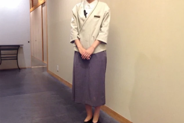 レストランの制服を着た女性。ベージュの着物のような上着に、グレーのロングスカートを着用している。