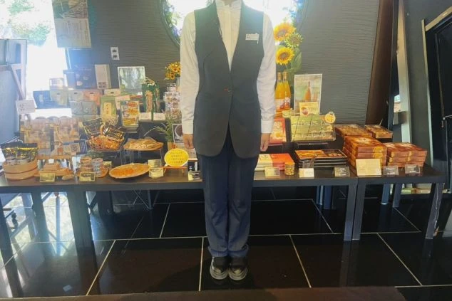黒いベストとパンツ、白い長袖シャツを着用したスタッフが、お土産が並んだ棚の前に立っている画像。リゾートバイトの仕事内容を表す、レストランまたは売店スタッフの制服の画像。