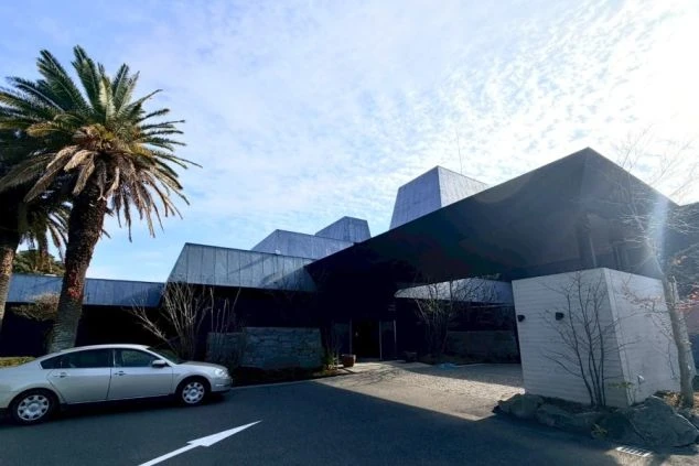 エントランスと建物外観。灰色のピラミッド型の屋根と、ベージュ色の壁、そして黒っぽい車寄せの庇（ひさし）が特徴的な建物。敷地内にはヤシの木が植えられている。