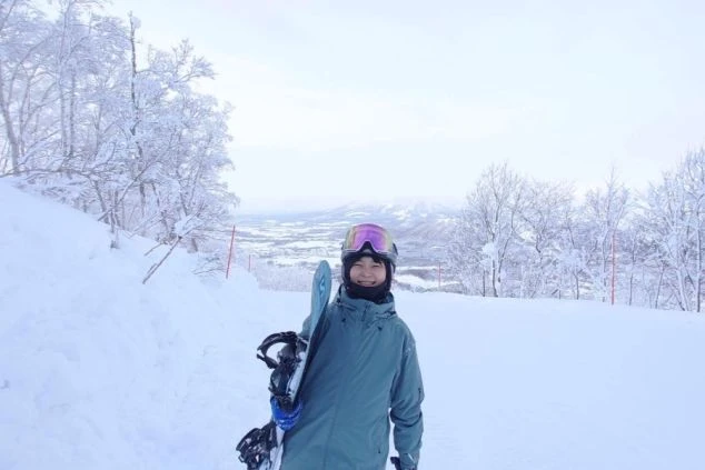 ワクトリでリゾートバイトをしたスタッフが撮影した、スノーボードを持ってカメラに笑顔を向ける女性。背景には雪景色が広がる。