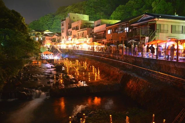 夜の温泉街。川には灯篭のような明かりが並び、川沿いには多くの屋台と人々で賑わっている。