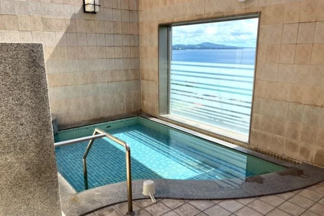 ワクトリの社員が出張で撮影した、リゾートバイトの寮にある大浴場の画像。窓の外には海が見え、タイル張りの浴槽には青いお湯が張られている。
