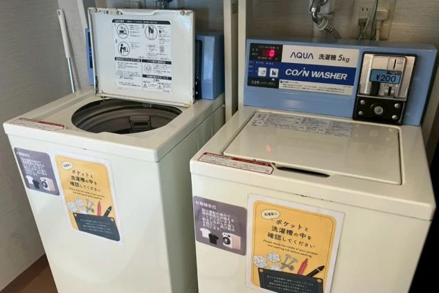 ワクトリの社員が出張で撮影した、リゾートバイトの寮に設置されたコインランドリーの洗濯機の画像。2台の縦型洗濯機が並び、使用方法の案内や料金が表示されている。