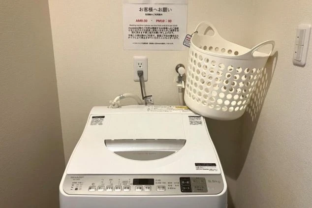 洗濯機と洗濯かごが置かれたランドリースペース。壁には「お客様へお願い」と書かれた利用案内の張り紙がある。