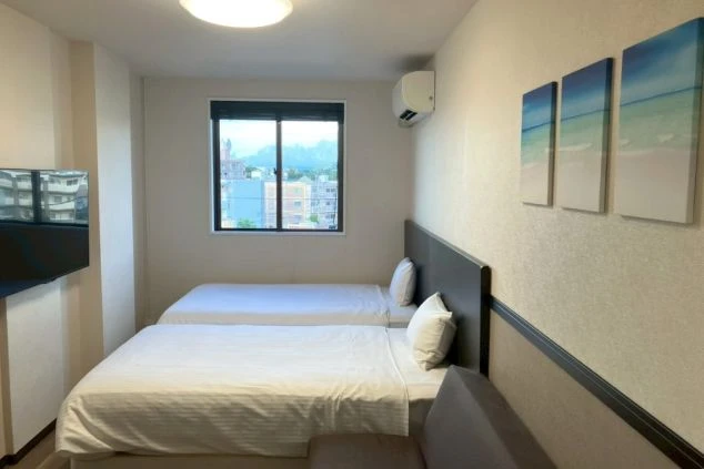 2つのベッドがあるシンプルなホテルの客室。部屋にはテレビ、エアコン、そして海をモチーフにしたアートが飾られている。