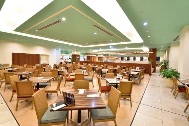  照明が明るいレストラン。複数のテーブルと椅子が規則正しく並び、奥には料理が並んでいる。