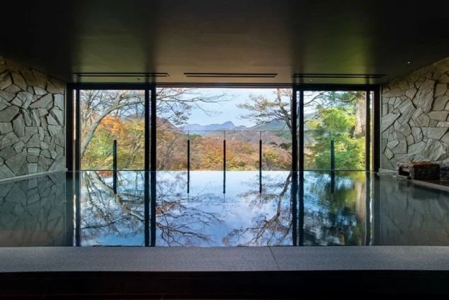 窓から紅葉した景色が見える、岩造りの内風呂。水面には木々が反射している。