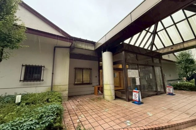 建物のエントランス(入口)。レンガ調の敷石が敷かれた地面と、ガラス張りの自動ドア、白い柱、そして屋根付きの通路が見える。