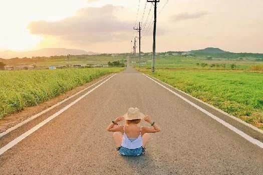 夕暮れ時、小浜島のシュガーロードで麦わら帽子をかぶった女性が座っている後ろ姿の画像。小浜島でのリゾートバイトの休日にのどかな道を散策するイメージ