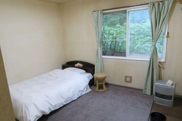 リゾートバイトの寮の部屋の中を撮影した画像。窓際にはヒーターが置かれており、窓の外には木々が見える。白く清潔な布団が敷かれたベッドと、小さなテーブルが配置された、一人暮らし用の個室の画像。