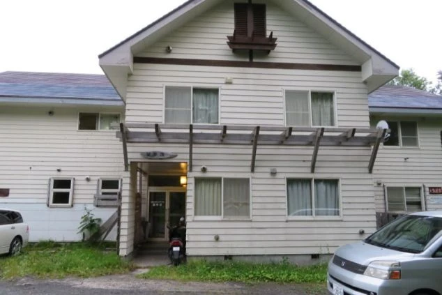 白壁が特徴的な2階建ての建物全体を捉えた画像。玄関口には屋根と木製のひさしがあり、車が2台駐車されている。リゾートバイトの寮として利用される、生活感のあるアパートのような外観の画像。