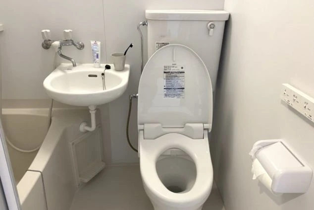 キッチンエリアが写っています。左側には銀色の冷蔵庫が見え、その右隣には白いカウンターとガスコンロがあります。コンロの上には複数の黒いバーナーが並んでいます。右奥には別の部屋へと続く通路が見えます。