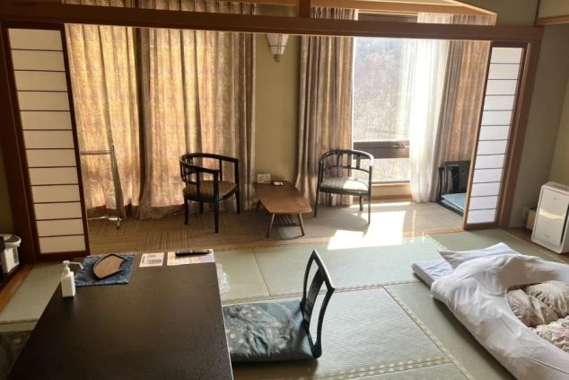 和室の寮の部屋の画像。畳敷きの空間に布団が敷かれ、座卓と椅子、窓際には椅子とテーブルがある。