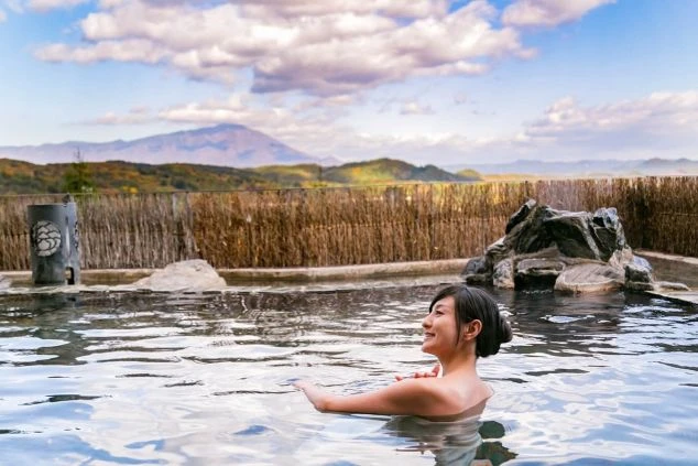 リゾートバイトの休日に温泉を楽しむイメージ画像。山々が見える開放的な露天風呂で、笑顔の女性が湯船につかっている。