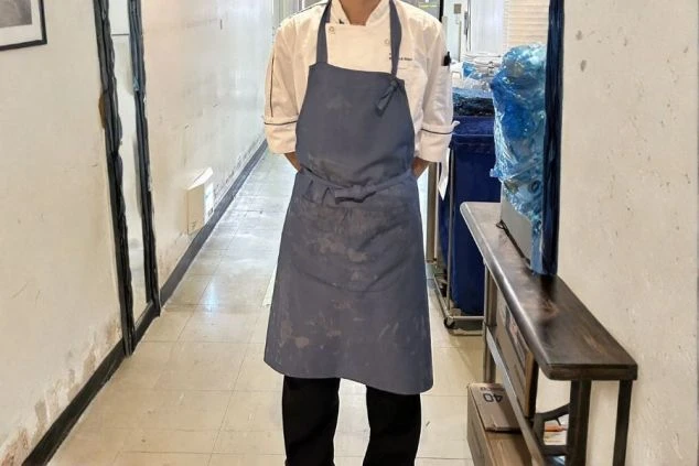 調理補助スタッフの制服画像。清潔感のある白のコックコートに、落ち着いたブルーのエプロンを合わせたスタイル。