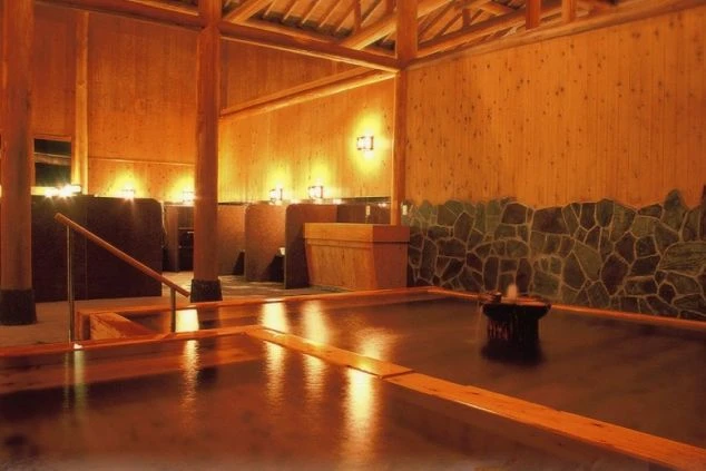木材をふんだんに使用した、温かい照明に照らされた和風の大浴場（温泉）。木組みの天井と壁、そして石垣調の壁を背景に、広々とした湯船が広がる。