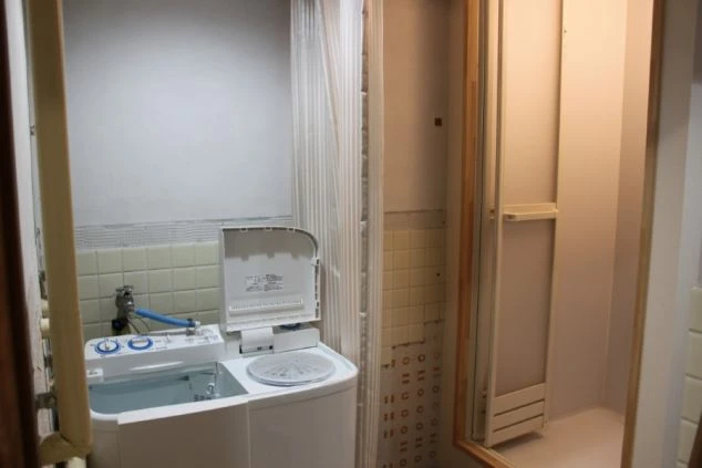 リゾートバイトのシェアタイプ個室寮の共同洗濯機の画像。洗濯機は二層式。右にはシャワーブースが見える。