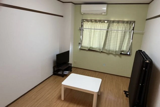 リゾートバイトの寮の個室。フローリングの部屋に、エアコン、テレビ、テーブル、折り畳みベッドが備え付けられている。