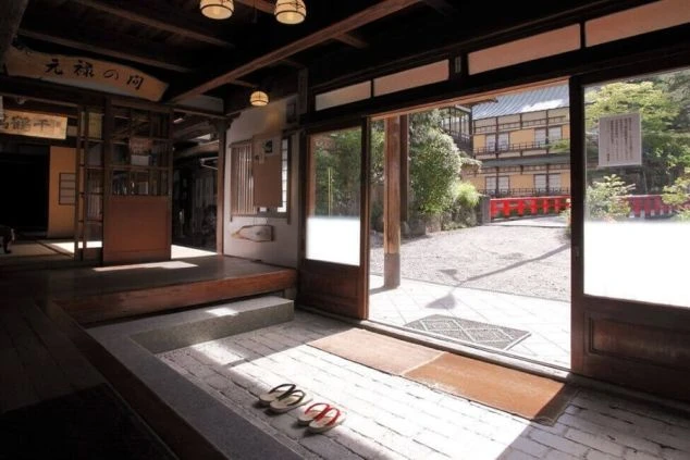 歴史を感じる古い木造建築の玄関。開放された引き戸からは、砂利の敷かれた中庭と赤い欄干の橋が見える。土間には草履が揃えられ、静かな和の風情が漂う空間。