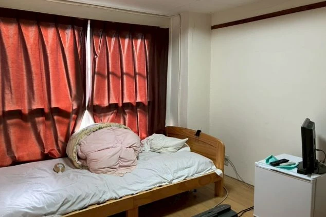 寮の居室。フローリングの床に木製フレームのベッドと布団が置かれ、窓には濃い赤のカーテンがかかっている。小型テレビとミニ冷蔵庫が備え付けられている。