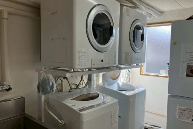 寮の共有家電スペース。青と白のチェック柄の布がかけられた台の上に、炊飯器、電子レンジ、そして食器類やラップなどが置かれている。