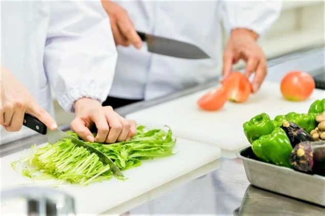 リゾートホテルの厨房で、白衣を着たスタッフ2名が水菜やトマトなどの新鮮な野菜を仕込んでいる様子。未経験から挑戦できるリゾートバイトの調理補助のイメージ画像。