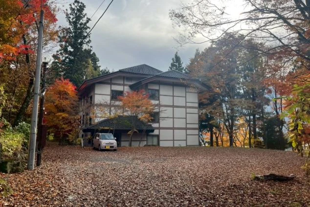 晩秋の時期と思われる、旅館の従業員寮または別館。白壁と茶色の木枠が特徴的な和洋折衷の建物が中央にあり、周囲は赤や黄色に色づいた木々と、地面を覆う落ち葉に囲まれている。手前には乗用車が一台停まっている。